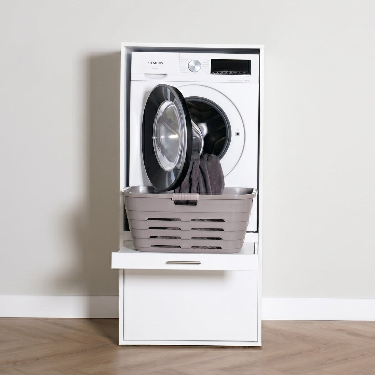 White front-loading washing machine with open door and dark laundry inside, placed on a white base cabinet with a drawer,.