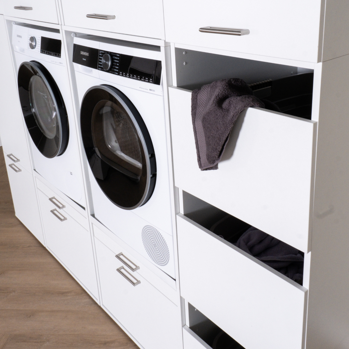 White built-in laundry cabinets with front-load washer and dryer, featuring open drawers with dark gray towels inside.