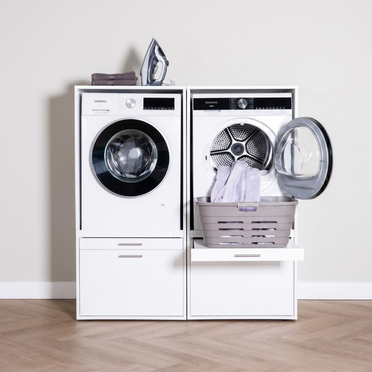White front-load washer and dryer set with laundry basket on open dryer door and iron and folded clothes on top.