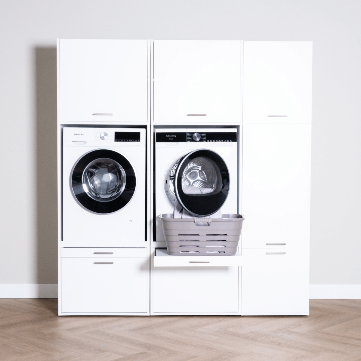 White built-in laundry unit with front-loading washer, dryer, and pull-out laundry basket on wooden floor.