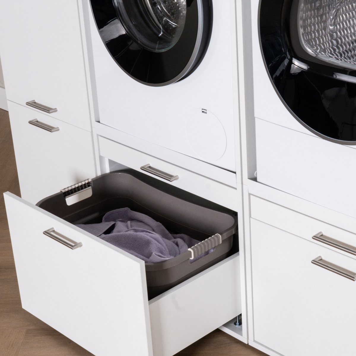 White built-in laundry cabinetry with front-loading washer and dryer, open drawer with dark gray laundry basket and folded.