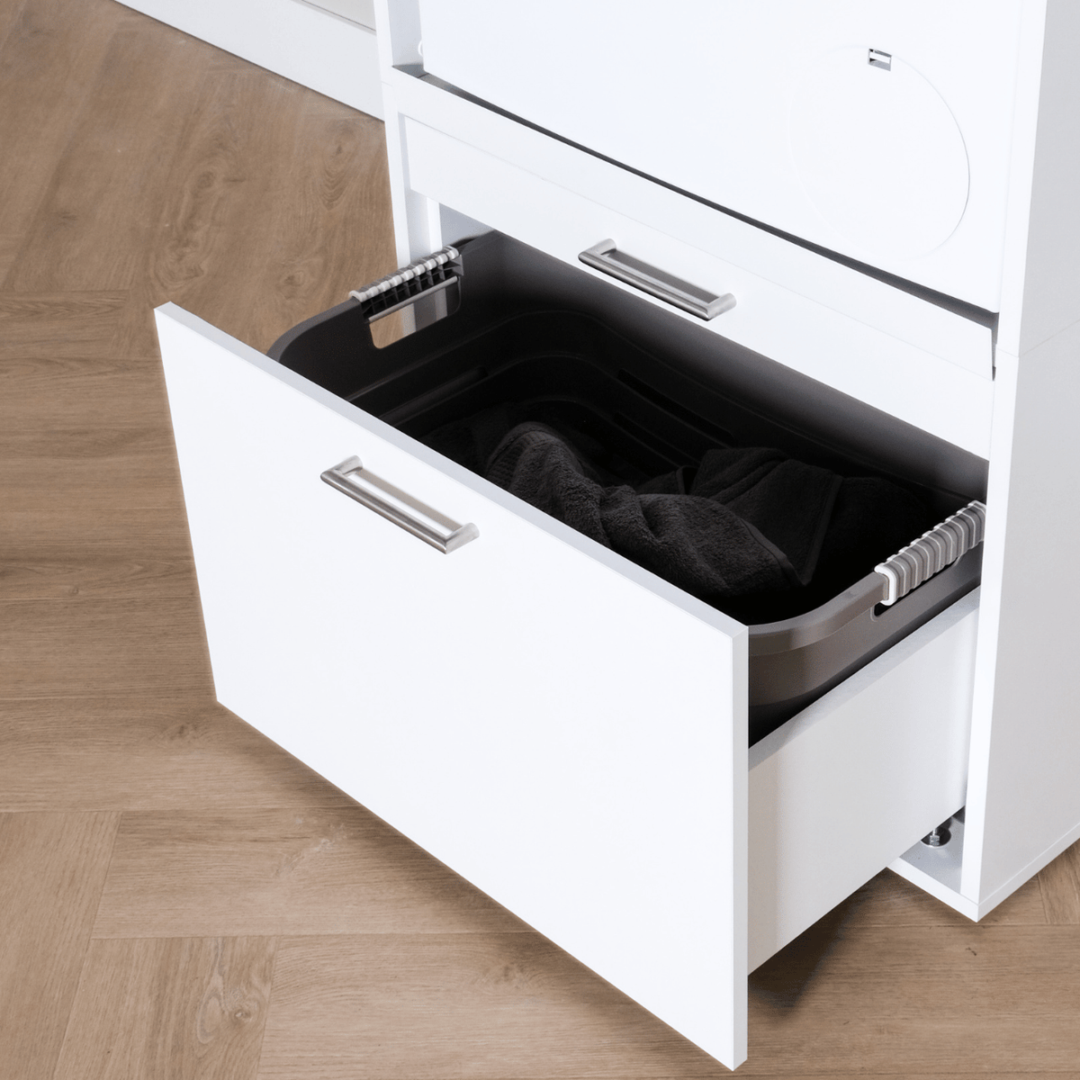 White modern cabinet with a pull-out drawer containing a black laundry basket and dark clothes inside.